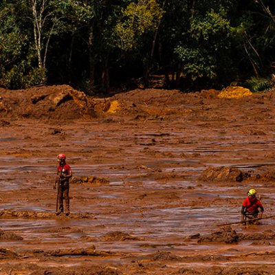 Two people in red shirts and helmets stand in red mud, there are trees in the background